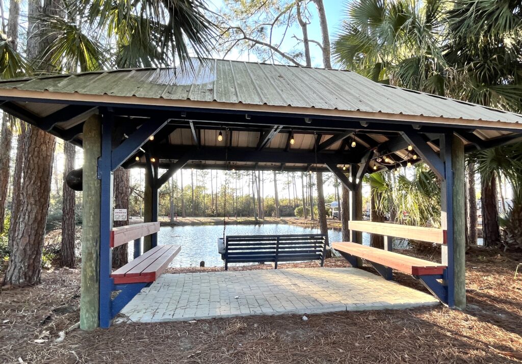 Large metal roof covered cabana with porch swing and two benches on either side, overlooking a pristine pond